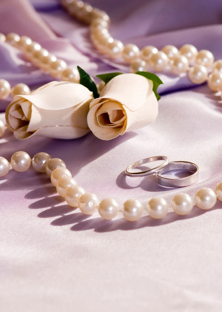 Pearls, white roses, and wedding rings rest on soft lilac satin with romantic elegance throughout.