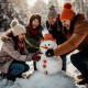 Photograph of a group of friends building a snowman on a snowy field, laughing together as they place a carrot for the nose