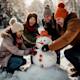 Photograph of a group of friends building a snowman on a snowy field, laughing together as they place a carrot for the nose