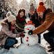 Photograph of a group of friends building a snowman on a snowy field, laughing together as they place a carrot for the nose