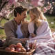 Photograph of a couple enjoying a romantic picnic, with a cozy blanket, basket of goodies, and surrounding soft pink flowers