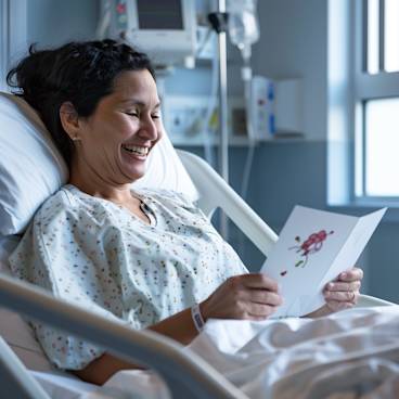 Photograph of a woman in a clean hospital bed smiling while reading a greeting card